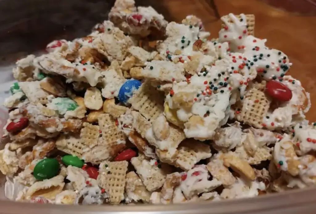 A bowl of White Trash dessert mix, with visible cereals, pretzels, nuts, and white chocolate, placed on a wooden table.