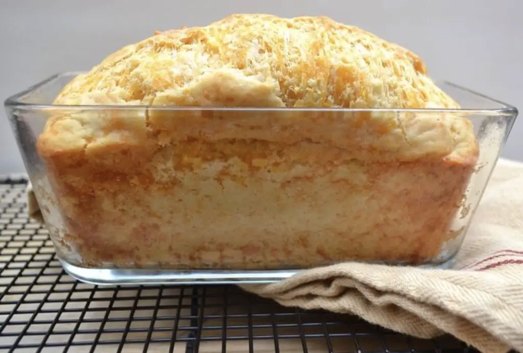 An inviting image of a golden-brown Cheddar Cheese Quick Bread loaf, with visible shreds of sharp cheddar, sliced and displayed on a wooden cutting board. A tempting treat that promises a moist and flavorful bite.