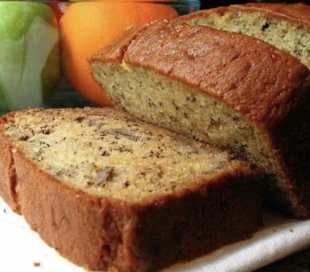 A golden-brown loaf of rich banana bread, adorned with slices of fresh bananas and a sprinkle of nuts, sits on a rustic wooden surface.
