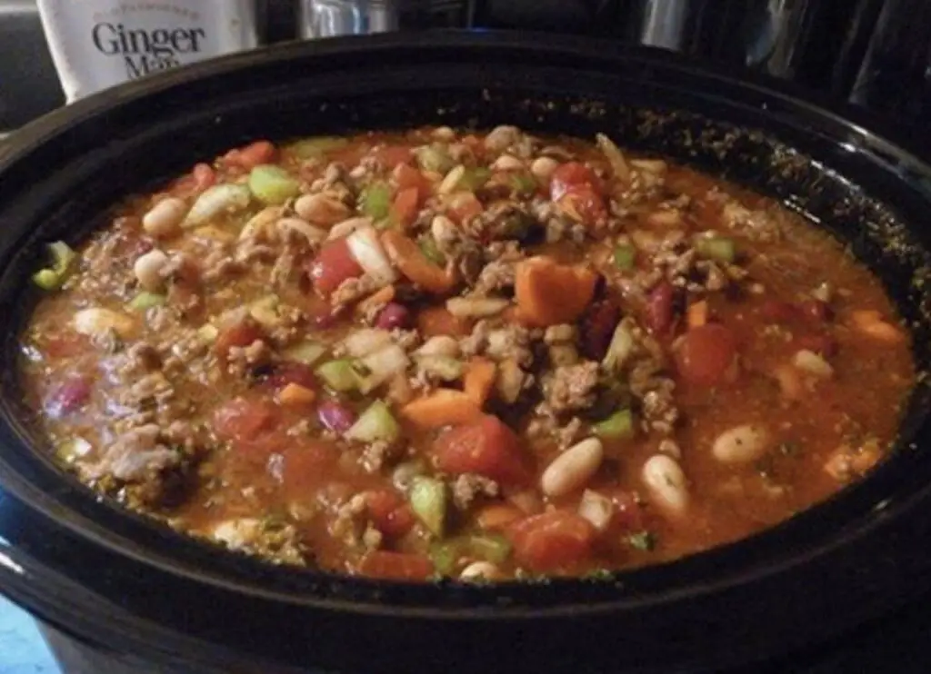 A steaming bowl of Hearty Crockpot Cowboy Soup, featuring chunks of beef, smoked sausage, beans, and veggies, garnished with melted cheddar, sour cream, and fresh green onions. Western comfort in every spoonful.