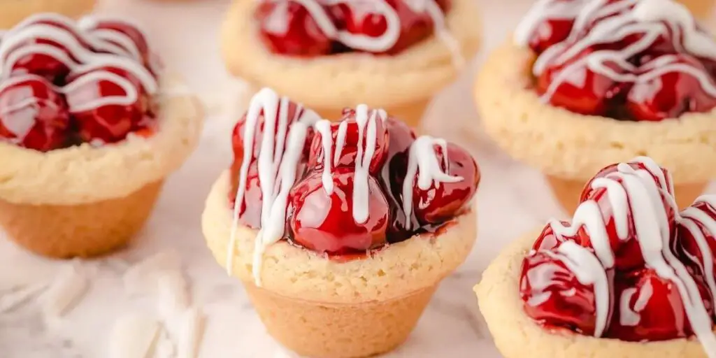 A batch of Cherry Pie Cookie Cups, featuring golden-brown, buttery crusts cradling sweet cherry pie filling, served in a mini muffin tin. A delightful, bite-sized treat!